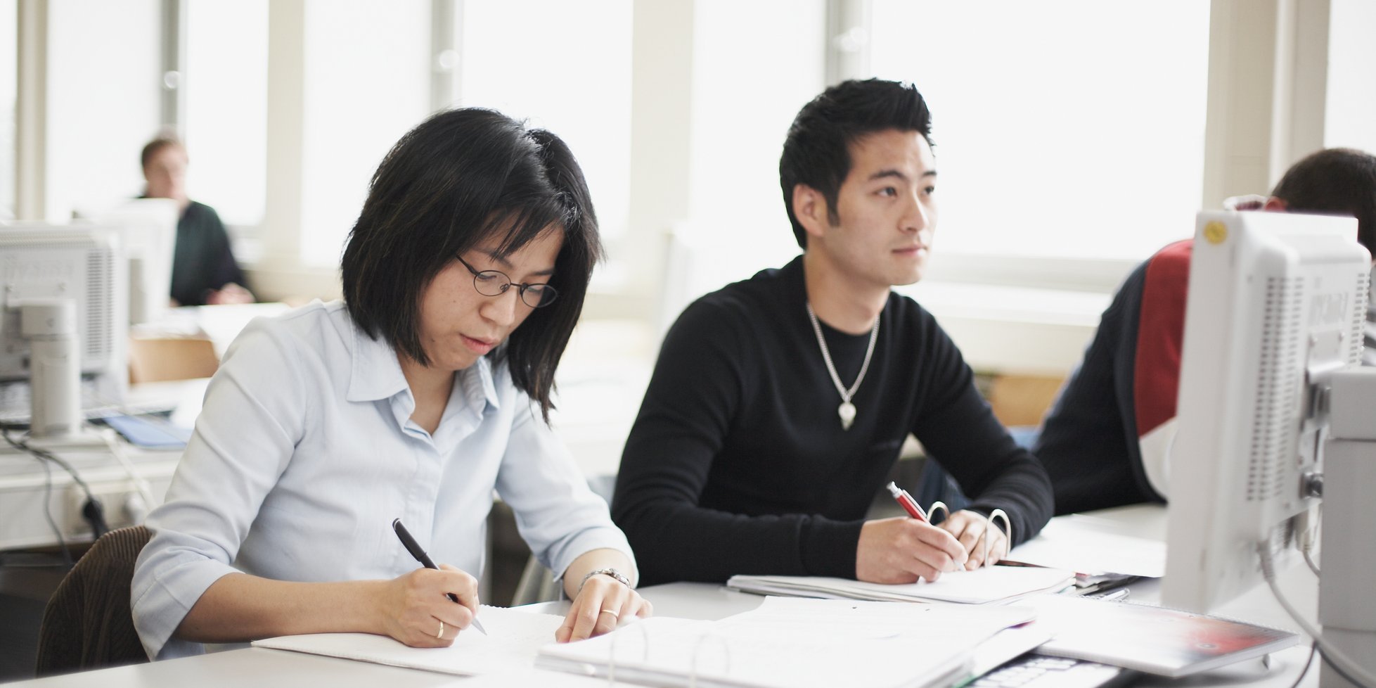 People sitting next to each other and writing at a table with computer monitor